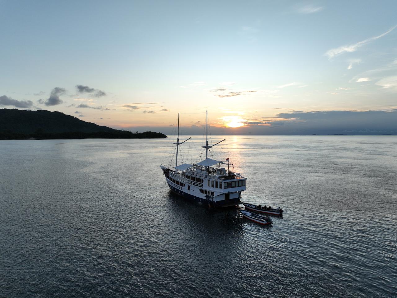 Pearl of Papua liveaboard anchored on calm waters at sunset near a lush island, with small boats tethered to the back, showcasing a serene maritime scene in Papua.