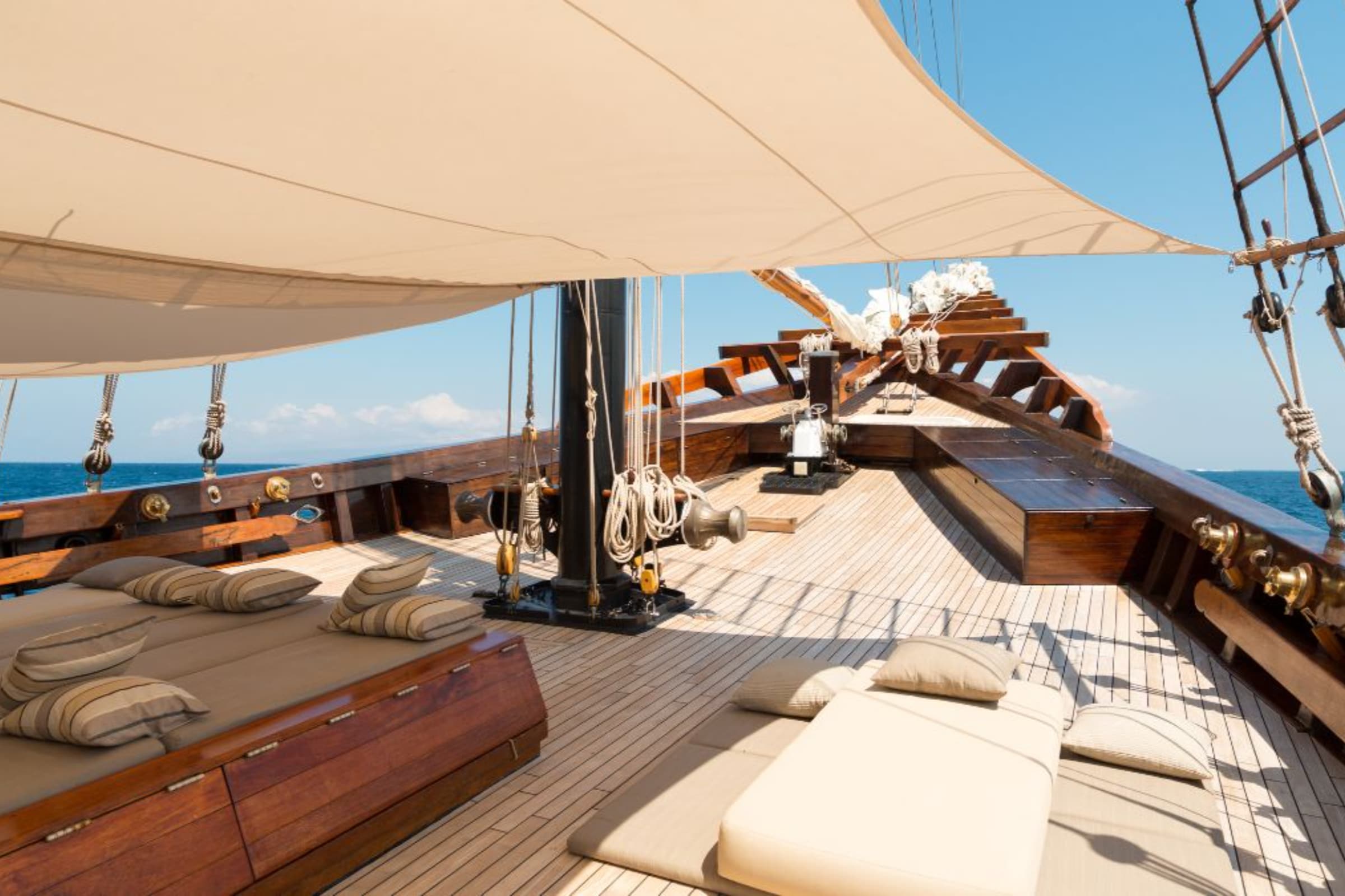 Luxury phinisi cruise Indonesia featuring a spacious wooden deck with comfortable lounge seating, elegant sails, and panoramic ocean views under a clear blue sky. Select 76 more words to run Humanizer.