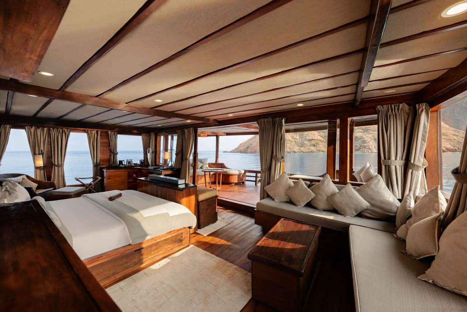 Luxury master cabin interior on a phinisi cruise Indonesia, featuring elegant wooden design, ocean-view windows, private seating area, and an open deck overlooking calm tropical waters